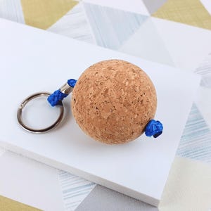 May include: A spherical cork keychain featuring a bright blue nylon cord threaded through the center. The cord connects to a circular silver-toned metal split ring. The natural tan cork has a textured, porous surface and a round, ball-like shape.