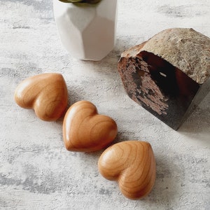 Può includere: Tre ornamenti in legno a forma di cuore in tonalità marroni, disposti in fila su una superficie bianca testurizzata.