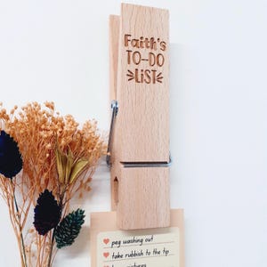 May include: A wooden clothespin with the text "Faith's TO-DO List" engraved on it. The clothespin is holding a brown card with a handwritten to-do list that includes "peg washing out" and "take rubbish to the tip".