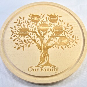 Op de afbeelding: Een rond houten snijplank met een gegraveerd boomontwerp. De boom heeft takken met bladeren en zes cirkels met namen erop: Mike, Janet, Simon, Jason, Sarah en Hugh. De tekst "Our Family" is gegraveerd aan de onderkant van de plank.