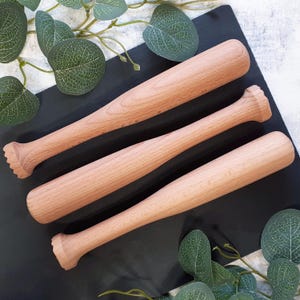 May include: Three wooden muddlers, also known as cocktail muddlers, are arranged on a black surface. The muddlers are made of light brown wood and have a rounded, slightly tapered shape. They are designed for crushing ingredients in cocktails.