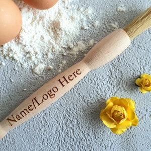 May include: A wooden pastry brush with natural bristles. The brush has the text "Name/Logo Here" engraved on the handle. The brush is lying on a gray surface with white flour and yellow flowers.