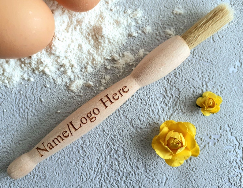 May include: A wooden pastry brush with natural bristles. The brush has the text "Name/Logo Here" engraved on the handle. The brush is lying on a gray surface with white flour and yellow flowers.