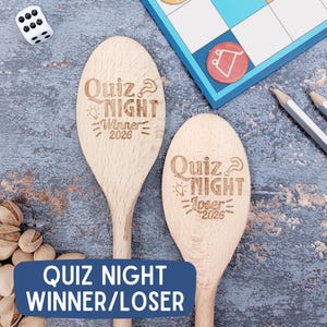 May include: Two wooden spoons engraved with "Quiz Night" and "Winner 2026" and "Loser 2026". A blue game board, pencils, and a die are in the background. The spoons are light brown and are on a gray surface. The text "Quiz Night Winner/Loser" is on a blue banner.