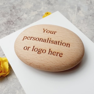 Może przedstawiać: Drewniana, owalna etykieta z wygrawerowanym tekstem "Your personalisation or logo here". Etykieta leży na białym tle.