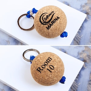 May include: A spherical cork keyring featuring a blue nylon cord and a silver metal ring. One side displays the black text Saltmere MARINA with a wave graphic, while the reverse side features the black text Room 10 printed on the cork surface.