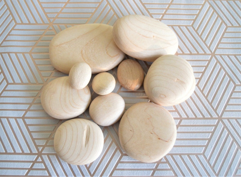 Puede incluir: Una colecci&oacute;n de piedras lisas de madera marr&oacute;n claro, que se asemejan a piedras de r&iacute;o, dispuestas sobre una superficie con un patr&oacute;n blanco y dorado.
