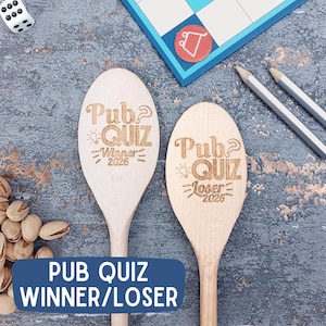 May include: Two wooden spoons with engraved text. One reads "Pub Quiz Winner 2026", the other "Pub Quiz Loser 2026". A blue banner at the bottom says "Pub Quiz Winner/Loser". Dice, pencils, and a game board are also visible.