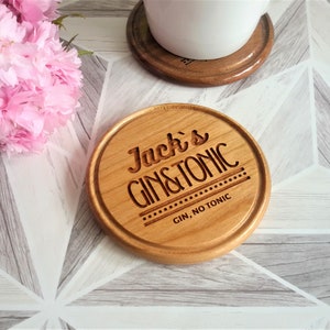 May include: A round wooden coaster with the text "Jack's Gin & Tonic" engraved on it. The coaster has a brown wood finish and a white background. The text is in a black font and is centred on the coaster. The coaster is sitting on a white and grey tiled surface.