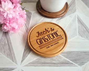 May include: A round wooden coaster with the text "Jack's Gin & Tonic" engraved on it. The coaster has a brown wood finish and a white background. The text is in a black font and is centered on the coaster. The coaster is sitting on a white and gray tiled surface.