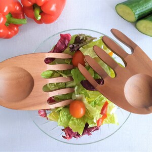 May include: Two wooden salad servers shaped like hands with a bowl on the back of each hand. The servers are holding a bowl of salad with red and green lettuce, cherry tomatoes, and red bell pepper.