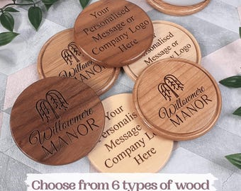 May include: A collection of circular wooden coasters in various wood tones, featuring laser-engraved text including Willowmere Manor and placeholders for custom messages or logos. The text Choose from 6 types of wood is visible.
