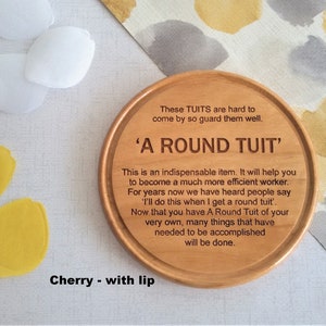 Puede incluir: Un posavasos redondo de madera con el texto "These TUITS are hard to come by so guard them well. 'A ROUND TUIT' This is an indispensable item. It will help you to become a much more efficient worker. For years now we have heard people say 'I'll do this when I get a round tuit.' Now that you have A Round Tuit of your very own, many things that have needed to be accomplished will be done." El posavasos está sobre una superficie blanca con flores de papel amarillas y blancas.