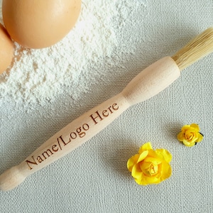 May include: A wooden pastry brush with a natural bristle brush head. The handle is engraved with the text "Name/Logo Here". The brush is on a white surface with two yellow flowers and two eggs.