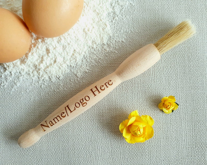 May include: A wooden pastry brush with a natural bristle brush head. The handle is engraved with the text "Name/Logo Here". The brush is on a white surface with two yellow flowers and two eggs.