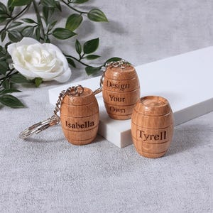 May include: Three small wooden barrel-shaped ornaments with metal hardware. Two barrels feature engraved names, Isabella and Tyrell, while the third barrel is engraved with the text Design Your Own. The barrels have a light brown wood grain finish.