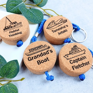 Può includere: Quattro portachiavi rotondi in sughero con cordini intrecciati blu e anelli in metallo. Ognuno presenta un'illustrazione incisa al laser di un'imbarcazione e il testo: Fishful Thinking, Tanesha's Jetski, Grandad's Boat e Captain Fletcher.