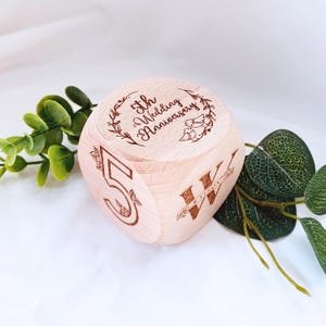 May include: A wooden cube with the text "5th Wedding Anniversary" engraved on one side, surrounded by a wreath and two birds. The number "5" is engraved on another side of the cube.