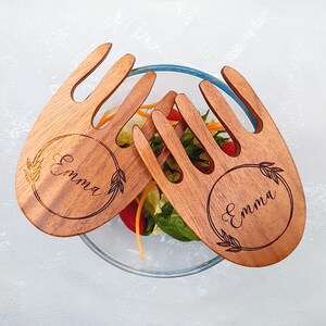 Puede incluir: Dos utensilios de ensalada de madera con un diseño en forma de tenedor. Cada utensilio tiene un grabado circular con el nombre "Emma" y elementos decorativos. Los utensilios descansan sobre un cuenco de vidrio lleno de ensalada.
