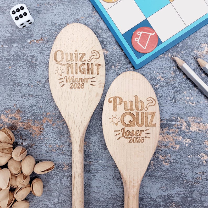 May include: Two wooden spoons with engraved text. One reads "Quiz Night Winner 2026" and the other "Pub Quiz Loser 2026". A dice, board game, pencils, and pistachios are also visible.