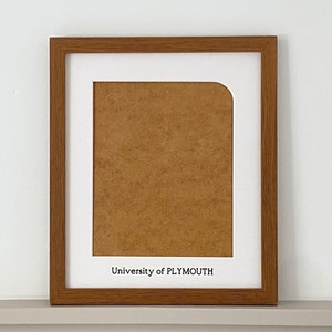 May include: A rectangular picture frame with a medium brown wood grain finish and a white mat board. The mat features a rounded top right corner and the black printed text University of PLYMOUTH centred at the bottom edge of the white border.