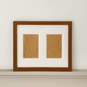 May include: A rectangular picture frame with a medium brown wood grain finish. The frame features a white mount with two vertical rectangular apertures, each containing a light tan backing board for displaying two separate photographs.