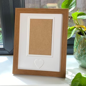 May include: A rectangular photo frame with a light brown wooden surround. It features a white mount with a heart-shaped embossed detail. The inner section is light brown. The frame is on a white surface.