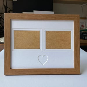 May include: A wooden picture frame with a white mount and two openings for photos. The mount has a heart-shaped cutout in the centre.