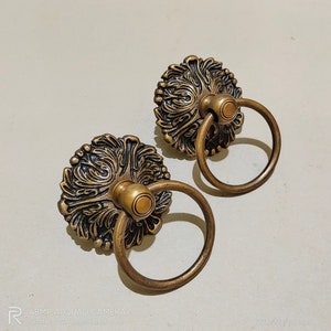 May include: Two ornate gold-toned cabinet door pulls with a ring handle. The pulls have a detailed floral design.
