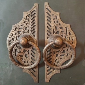 Może przedstawiać: Para ozdobnych, mosiężnych klamki do drzwi z misternymi wzorami kwiatowymi i uchwytami w kształcie pierścieni. Klamki są zamontowane na ciemnoszarym tle.