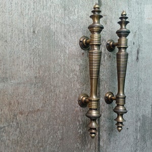 Op de afbeelding: Twee antieke messing deurgrepen met een decoratief, sierlijk ontwerp. De grepen zijn lang en slank met een puntige bovenkant en een geribbeld lichaam.