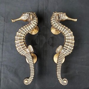 Peut inclure: Deux poign&eacute;es de porte en bronze en forme d'hippocampe. Les poign&eacute;es pr&eacute;sentent des &eacute;cailles et des nageoires d&eacute;taill&eacute;es, mont&eacute;es sur des plaques arri&egrave;re rondes en laiton. Les hippocampes courbent gracieusement, ajoutant une touche nautique unique.