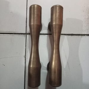 May include: A pair of vertical door handles made of brushed brass with a smooth, elongated, and slightly hourglass-shaped silhouette. Each handle features a rounded top and bottom with a tapered center, providing a consistent metallic finish.