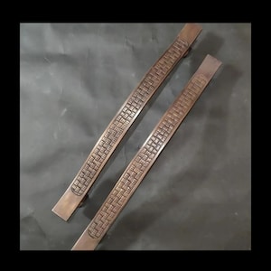 May include: Two long, bronze door handles with a woven pattern. The handles are curved and have a smooth, polished finish.