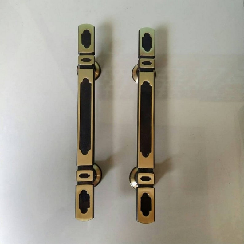May include: Two gold door handles with black accents and intricate designs. The handles are rectangular with a rounded top and bottom.