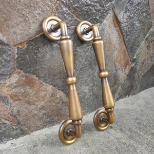 May include: A pair of antique brass door handles. Each handle has a circular backplate, a decorative central section, and a curved handle. The handles are made of brass and have a warm, aged finish. These handles are designed for doors.