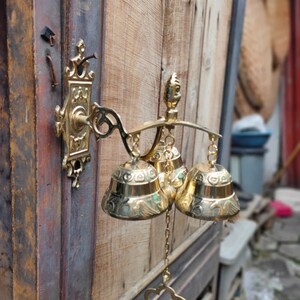 May include: A gold-toned metal door chime with three bells. The bells are decorated with intricate designs and hang from a decorative metal frame. The chime is attached to a wooden door.