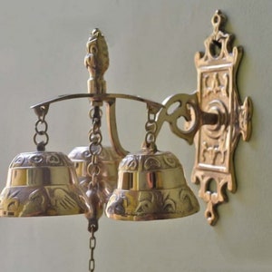 May include: A set of three brass bells with intricate designs hanging from a decorative wall mount. The bells are attached to a chain and have a polished finish.