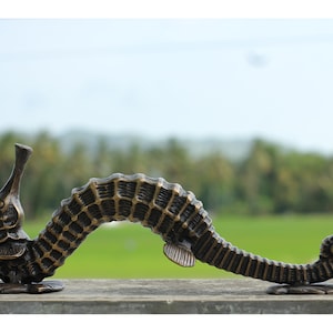 Peut inclure: Une sculpture de bronze repr&eacute;sentant un hippocampe avec des d&eacute;tails complexes. L'hippocampe est courb&eacute;, avec sa queue enroul&eacute;e. La sculpture est plac&eacute;e sur un fond flou de feuillage vert et de ciel bleu clair.
