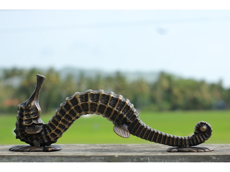 Peut inclure: Une sculpture de bronze repr&eacute;sentant un hippocampe avec des d&eacute;tails complexes. L'hippocampe est courb&eacute;, avec sa queue enroul&eacute;e. La sculpture est plac&eacute;e sur un fond flou de feuillage vert et de ciel bleu clair.