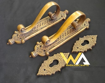 May include: Antique brass door hardware set featuring two ornate handles with curved grips and decorative backplates. Includes two keyhole covers. The set has an aged, vintage appearance, with intricate engraved patterns and a warm, golden hue.