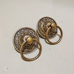 Puede incluir: Dos tiradores de puerta de latón adornados con intrincados diseños florales y asas de anillo. Los tiradores son de color dorado cálido y tienen una estética vintage.