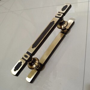 May include: A gold door handle with a black accent. The handle is made of metal and has a rectangular shape. It is designed to be used on a glass door.
