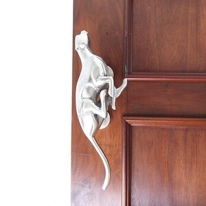 Könnte beinhalten: Ein silberner Türklopfer in Form eines Hundes, mit dem Kopf nach hinten geneigt und dem Schwanz eingerollt. Der Türklopfer ist an einer Holztür montiert.