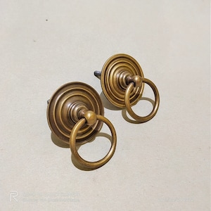 Può includere: Due pomelli per armadi in ottone antico con anelli di trazione. Ogni pomo ha una base circolare con anelli concentrici e un centro rialzato. Gli anelli sono attaccati a un anello metallico circolare.