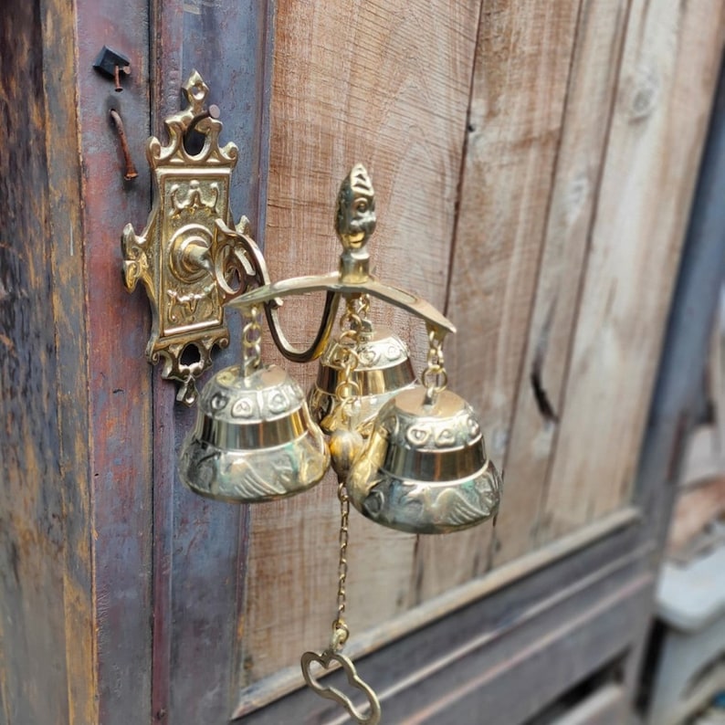 May include: A gold-toned metal door chime with three bells. The chime is mounted on a wooden door. The bells are decorated with intricate designs.