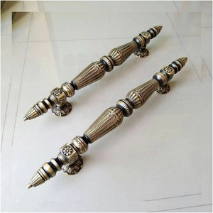 May include: Two ornate gold door handles with intricate details and a black finish. The handles are designed with a pointed tip and a decorative base.