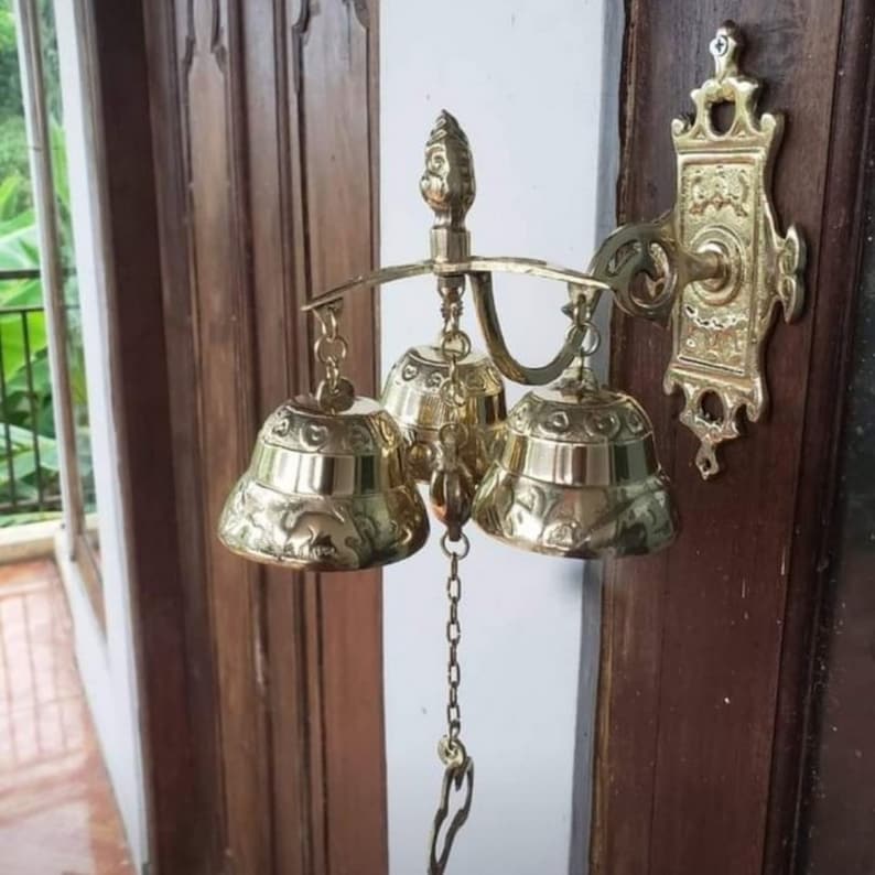 May include: A gold-toned triple bell door chime with a decorative ornate design. The bells are attached to a metal frame with a chain hanging from the center bell.