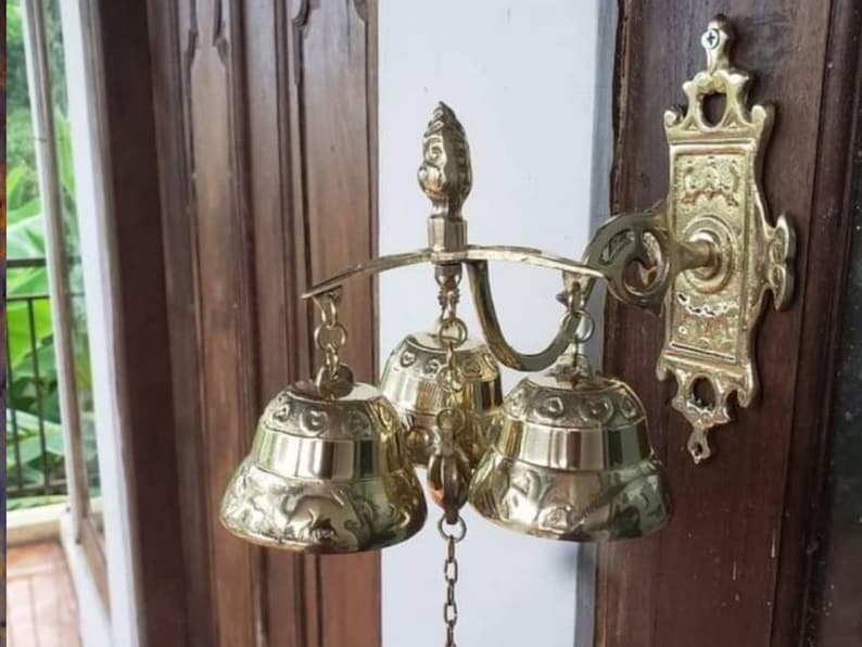 May include: A gold-toned metal door chime with three bells and a decorative wall mount. The bells are ornate and have a detailed design.