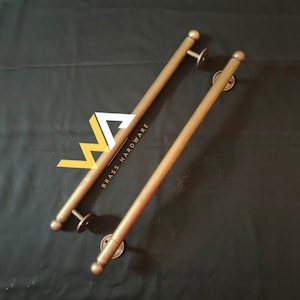 May include: Two long, gold-toned metal door handles with rounded ends. The handles are attached to round metal plates. The text "BRASS HARDWARE" is visible on one of the handles.